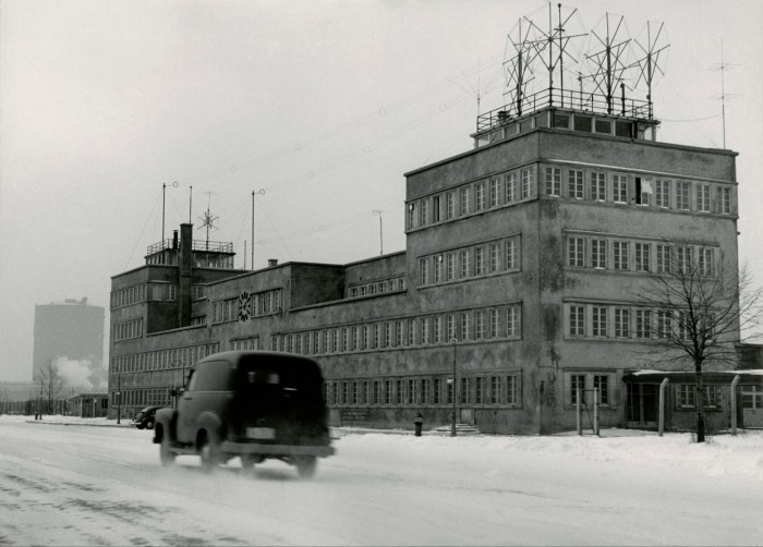A black and white photograph of an old car driving past a large square building with lots of antennas on the flat roof