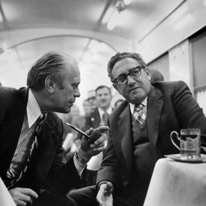 A black and white photograph of two men in suits leaning into one another. One man has a pipe in his hand. There are men behind them