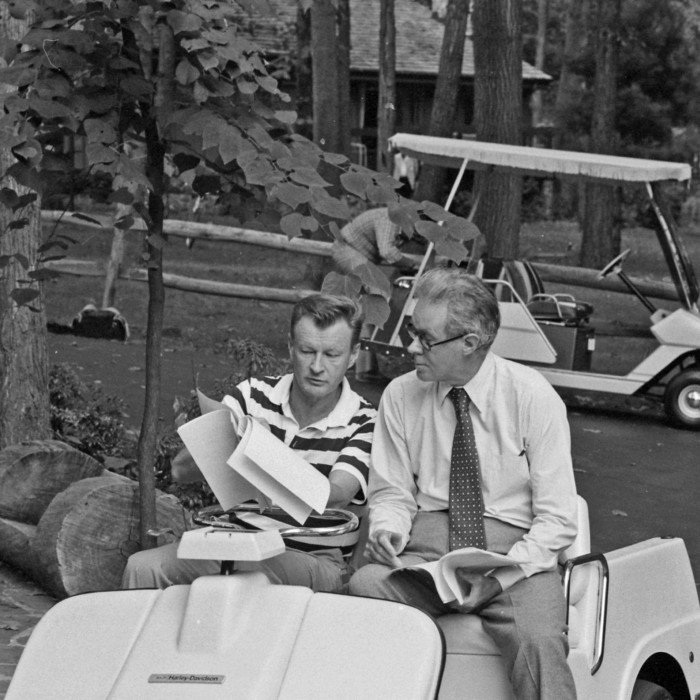 A black and white photograph of two men in a golf buggy looking over some documents 