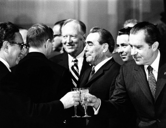 A black and white photograph of a group of men dressed in suits. Two of the men have small glasses in their hands and are toasting