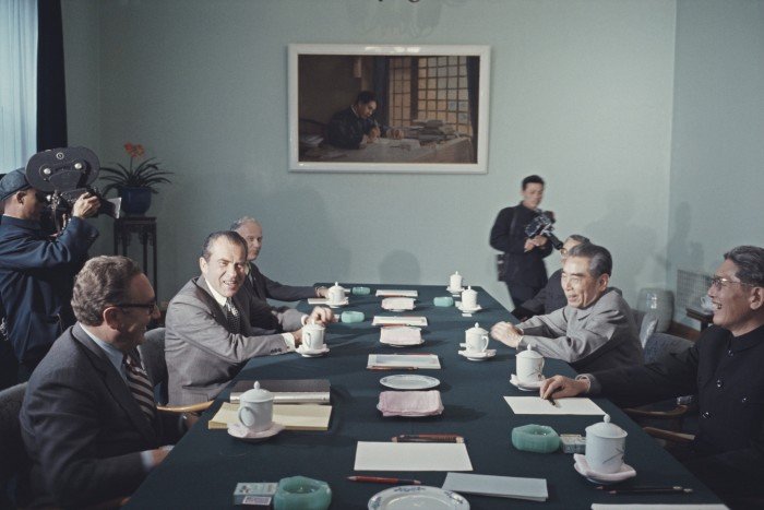 A group of men sitting around a table. There are two cameramen in the background