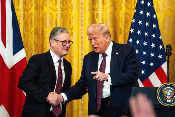 Two men in suits shake hands in front of a yellow curtain. There is an American flag on the right and a British flag on the left
