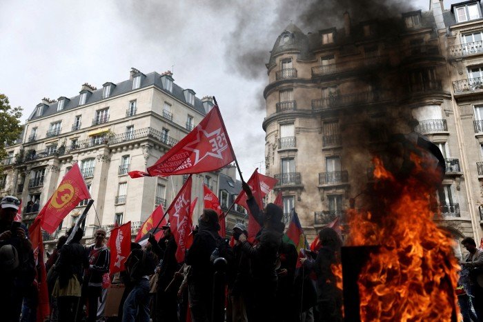 Protesters wave red flags near a burning garbage bin, with smoke rising in front of Parisian buildings during a demonstration.